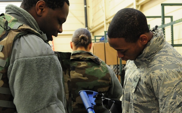 Two military personnel in the foreground are examining a blue training weapon (inert/dummy firearm), likely during a training exercise or equipment issue session. A third service member is visible in the background wearing woodland camouflage. The setting appears to be a military warehouse or supply facility with metal shelving visible.