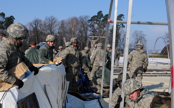 A large group of military personnel in camouflage uniforms are assembling or erecting a large tent or temporary shelter structure outdoors. The scene appears to take place in a cold, wintry environment with snow visible on the ground and bare trees in the background. Personnel from what appears to be the U.S. Air Force are working collaboratively on the construction task.