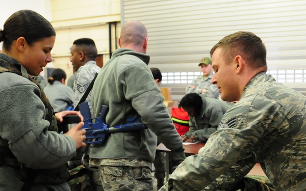 Military personnel, appearing to be U.S. Air Force members based on their Airman Battle Uniform (ABU) pattern, are engaged in an equipment issue or inspection session inside a warehouse or hangar facility. Several airmen are handling blue-colored training weapons and woodland camouflage tactical vests at a table. The scene suggests a pre-deployment readiness exercise or equipment draw event.