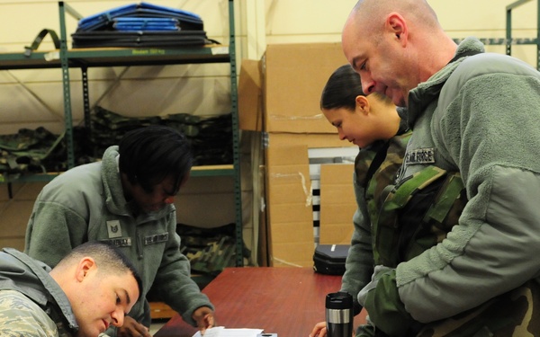 Military personnel, appearing to be U.S. Air Force members based on visible uniforms and insignia, are gathered in what appears to be a supply or equipment warehouse. Several individuals are filling out or reviewing paperwork at a wooden table, while others stand nearby. The setting suggests an equipment issue, accountability check, or administrative processing event.