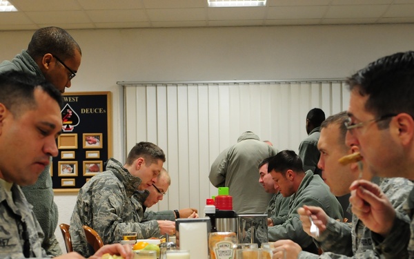 Military personnel in US Air Force Airman Battle Uniforms (ABU) are eating a meal together in what appears to be a military dining facility (DFAC/chow hall). Multiple service members are seated at tables with food trays containing breakfast items. The setting includes a bulletin board with photos and unit insignia visible on the wall in the background.