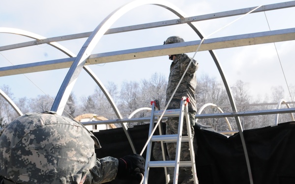Military personnel in camouflage uniforms are constructing or assembling a large tent or temporary shelter structure. One soldier stands on an aluminum step ladder working on the upper frame, while another in the foreground stabilizes guide ropes or cables. The setting appears to be an outdoor military installation during cold weather, with bare trees visible in the background.