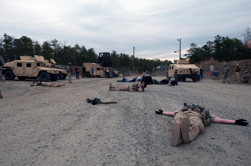 A military training exercise is underway on a gravel area, with multiple personnel in desert camouflage uniforms lying on the ground in simulated casualty positions. Several military vehicles including HMMWVs (Humvees) and a heavy truck are visible in the background. Standing personnel appear to be observers, instructors, or role-players overseeing the scenario.