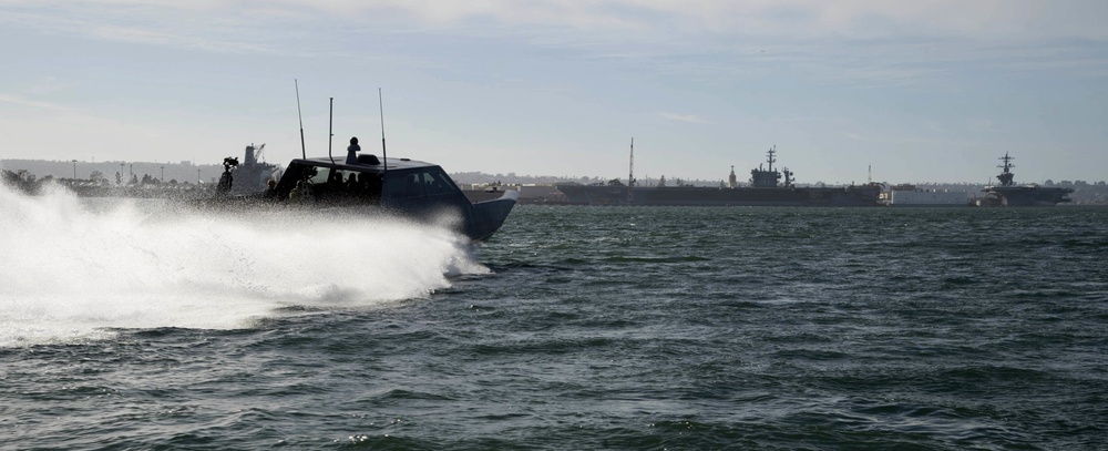 A high-speed military or tactical watercraft is moving rapidly across open water, generating a large white wake and spray. In the background, what appear to be large naval vessels, likely aircraft carriers or amphibious assault ships, are anchored or moored near a distant shoreline. The scene suggests a naval operational environment, possibly near a military base or harbor.