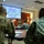 A military briefing or training session is taking place in a conference room, with a uniformed presenter standing at the front addressing seated military personnel. A projection screen displays what appears to be a website or portal, partially readable as 'MyVeHU' or similar. Attendees are wearing various camouflage uniforms and are seated at tables with documents.