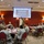 A military briefing or training session is taking place in a conference room with multiple soldiers in Army Combat Uniforms (ACU) seated at round tables. An instructor in uniform is pointing toward a projected presentation screen at the front of the room. The setting appears to be a formal military training or educational environment.