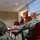 A civilian presenter in a dark suit is delivering a briefing at a podium to an audience that includes at least two military personnel in camouflage uniforms. The presentation slide visible on the projection screen is titled 'Today's Objectives' and references RCTP training topics. The setting appears to be a conference or training room with standard institutional furnishings.