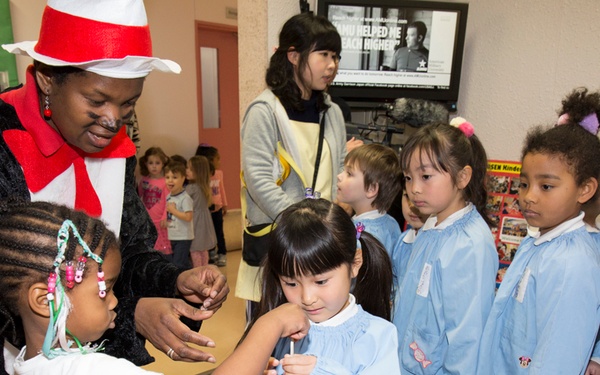 An adult dressed in a Cat in the Hat costume is interacting with a group of young children, likely during a Dr. Seuss-themed reading event. Most of the children are wearing matching light blue smocks, suggesting they are from the same school or class on a field trip. The setting appears to be a library or community center with Dr. Seuss decorations visible in the background.