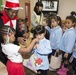 An adult dressed in a Cat in the Hat costume is interacting with a group of young children, likely during a Dr. Seuss-themed reading event. Most of the children are wearing matching light blue smocks, suggesting they are from the same school or class on a field trip. The setting appears to be a library or community center with Dr. Seuss decorations visible in the background.