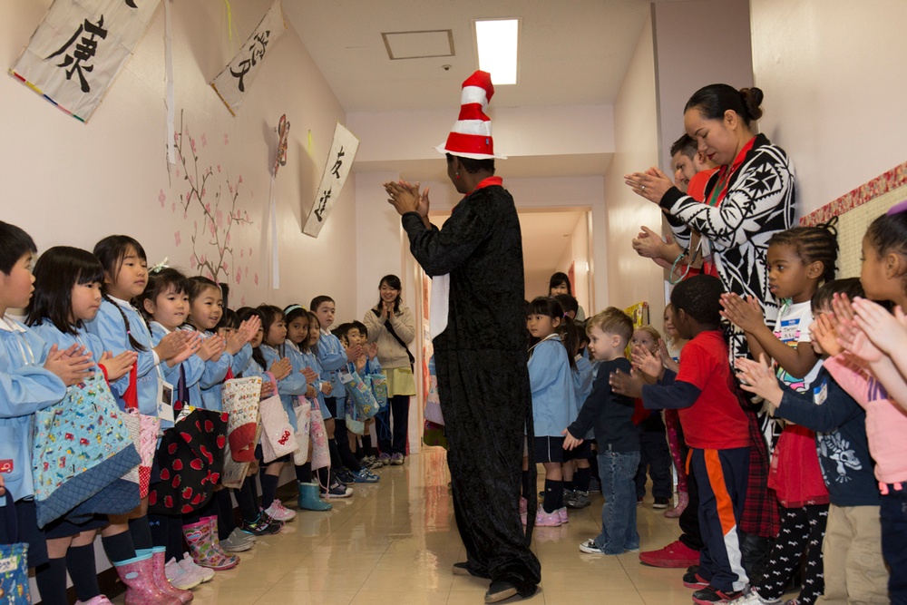 A person dressed in a Cat in the Hat costume (black outfit with red and white striped tall hat) walks through a corridor lined with clapping children and adults. The setting appears to be a Japanese school or kindergarten, evidenced by calligraphy banners on the walls, cherry blossom decorations, and children wearing light blue school uniforms. The event appears to be a multicultural gathering, with both Japanese children and children of other ethnicities present alongside adult supervisors.