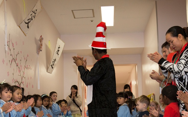 A person dressed in a Cat in the Hat costume (black outfit with red and white striped tall hat) walks through a corridor lined with clapping children and adults. The setting appears to be a Japanese school or kindergarten, evidenced by calligraphy banners on the walls, cherry blossom decorations, and children wearing light blue school uniforms. The event appears to be a multicultural gathering, with both Japanese children and children of other ethnicities present alongside adult supervisors.