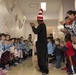 A person dressed in a Cat in the Hat costume (black outfit with red and white striped tall hat) walks through a corridor lined with clapping children and adults. The setting appears to be a Japanese school or kindergarten, evidenced by calligraphy banners on the walls, cherry blossom decorations, and children wearing light blue school uniforms. The event appears to be a multicultural gathering, with both Japanese children and children of other ethnicities present alongside adult supervisors.