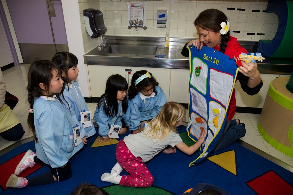 A female educator or museum guide is conducting a hands-on sorting activity with a group of young children, approximately 4-6 years old, in what appears to be a children's museum or educational facility. She is holding up a felt sorting board labeled 'Sort It!' while the children, most wearing matching light blue smocks with name tags, engage with the activity. One blonde child in pink polka-dot pants is actively reaching toward the board.