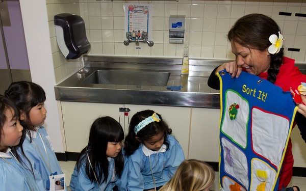 A female educator or museum guide is conducting a hands-on sorting activity with a group of young children, approximately 4-6 years old, in what appears to be a children's museum or educational facility. She is holding up a felt sorting board labeled 'Sort It!' while the children, most wearing matching light blue smocks with name tags, engage with the activity. One blonde child in pink polka-dot pants is actively reaching toward the board.