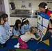 A female educator or museum guide is conducting a hands-on sorting activity with a group of young children, approximately 4-6 years old, in what appears to be a children's museum or educational facility. She is holding up a felt sorting board labeled 'Sort It!' while the children, most wearing matching light blue smocks with name tags, engage with the activity. One blonde child in pink polka-dot pants is actively reaching toward the board.