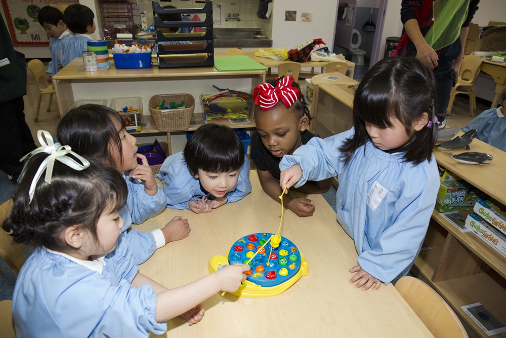 A group of five young children in a classroom setting are gathered around a table playing a toy fishing game. The children are wearing light blue smocks/aprons, suggesting a school or daycare environment. The classroom is well-equipped with various educational materials, shelves, and activity stations visible in the background.