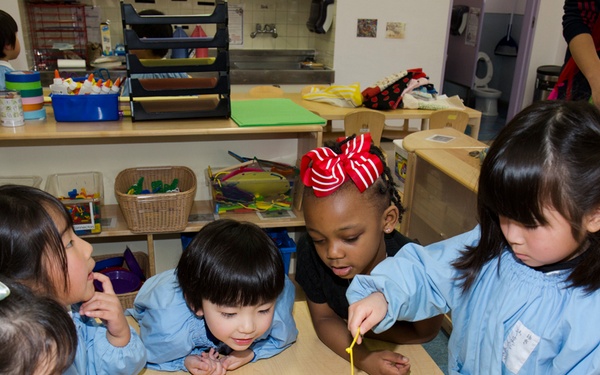A group of five young children in a classroom setting are gathered around a table playing a toy fishing game. The children are wearing light blue smocks/aprons, suggesting a school or daycare environment. The classroom is well-equipped with various educational materials, shelves, and activity stations visible in the background.