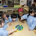 A group of five young children in a classroom setting are gathered around a table playing a toy fishing game. The children are wearing light blue smocks/aprons, suggesting a school or daycare environment. The classroom is well-equipped with various educational materials, shelves, and activity stations visible in the background.