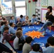A teacher is conducting a read-aloud session with a group of young elementary school students seated on a colorful blue rug featuring a sun design. The classroom setting appears to be a kindergarten or early primary classroom with educational posters, a behavior chart, and a 'School Days Number Chart' visible on the walls. The children, many wearing light blue uniforms, are attentively gathered around the teacher who is seated on a small chair holding an open book.