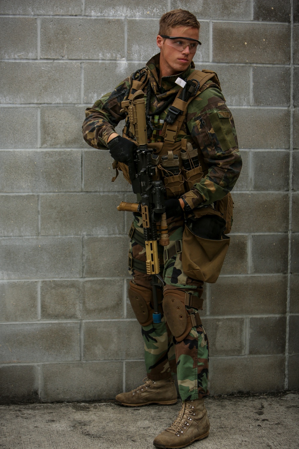 A young male individual is posed against a concrete block wall wearing woodland camouflage tactical gear and carrying an airsoft or replica firearm. The equipment includes a tactical vest, knee pads, gloves, protective eyewear, and a backpack. The setup is consistent with airsoft or milsim (military simulation) recreational activity.