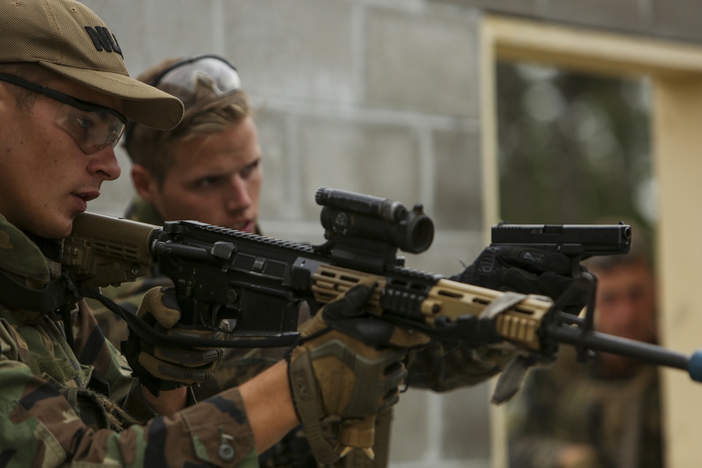 The image shows military or law enforcement personnel in camouflage uniforms conducting what appears to be a weapons training exercise. A primary subject in the foreground is aiming an AR-style rifle equipped with an optic sight and tactical accessories, while a second individual observes closely from behind. The setting appears to be an urban training environment near a concrete block structure.