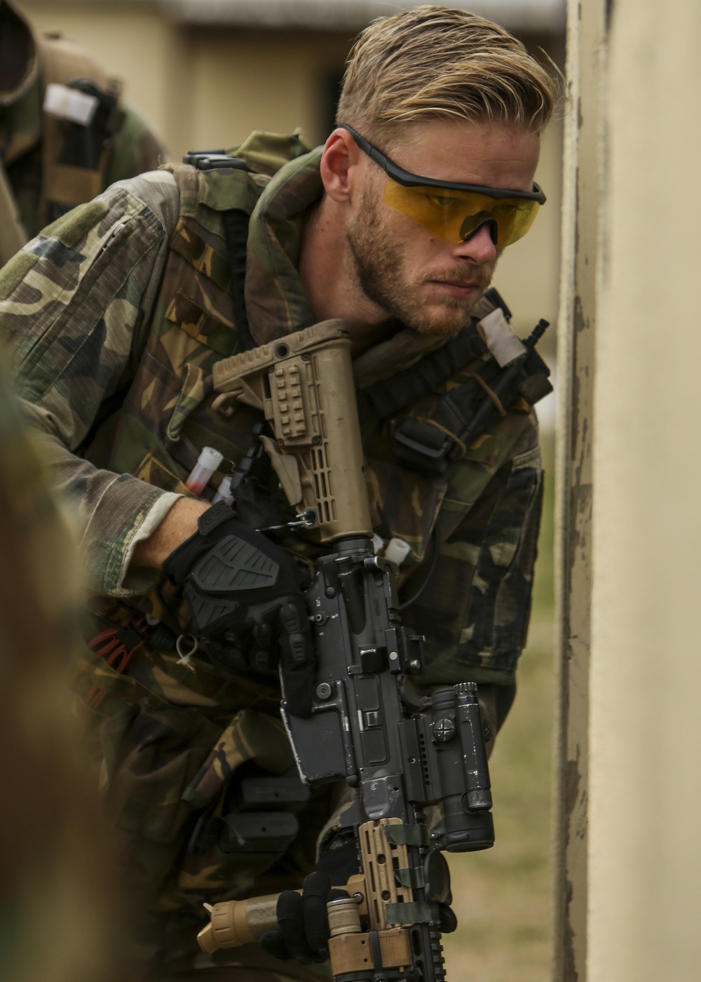 A male individual wearing woodland camouflage uniform and yellow-tinted protective eyewear is holding a tactical rifle with accessories. He appears to be in a tactical or training posture, leaning against a wall or doorframe. The setting suggests a military exercise, airsoft event, or tactical training scenario.