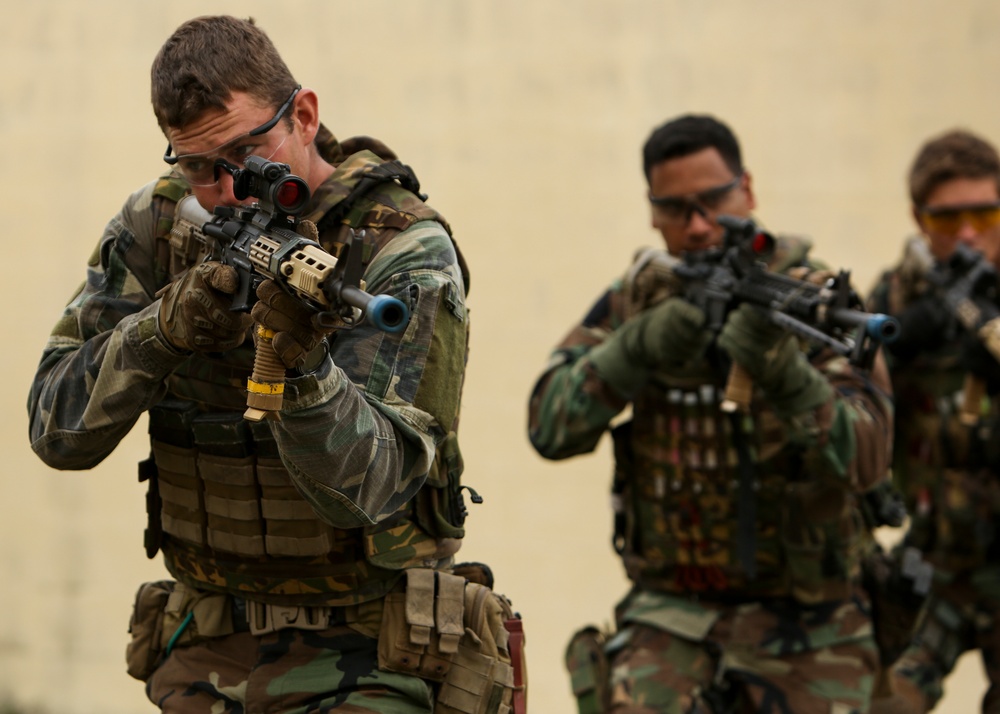 Three individuals in camouflage military-style uniforms and tactical vests are advancing in formation while aiming rifles with optic scopes. The foreground subject is in sharp focus, wearing gloves and protective eyewear pushed up on his forehead, while two others are visible in the background in similar postures. The setting appears to be a training exercise based on the controlled environment and safety-colored muzzle devices visible on the weapons.
