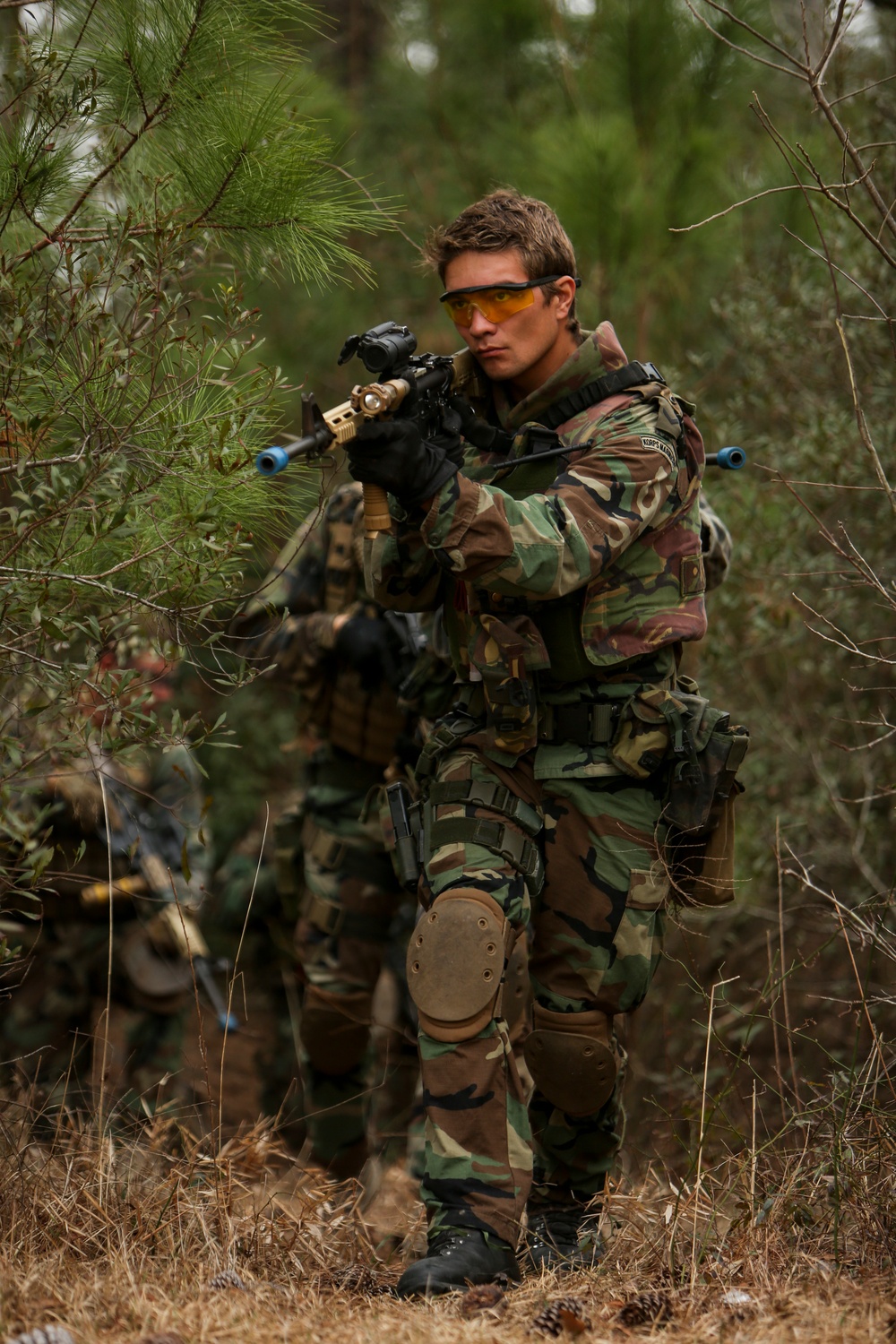 A person in woodland camouflage gear is aiming a replica airsoft rifle in an outdoor wooded setting. They are equipped with tactical gear including knee pads, a chest rig, and yellow-tinted protective eyewear. A second similarly equipped individual is visible in the background, suggesting a group airsoft or milsim (military simulation) activity.