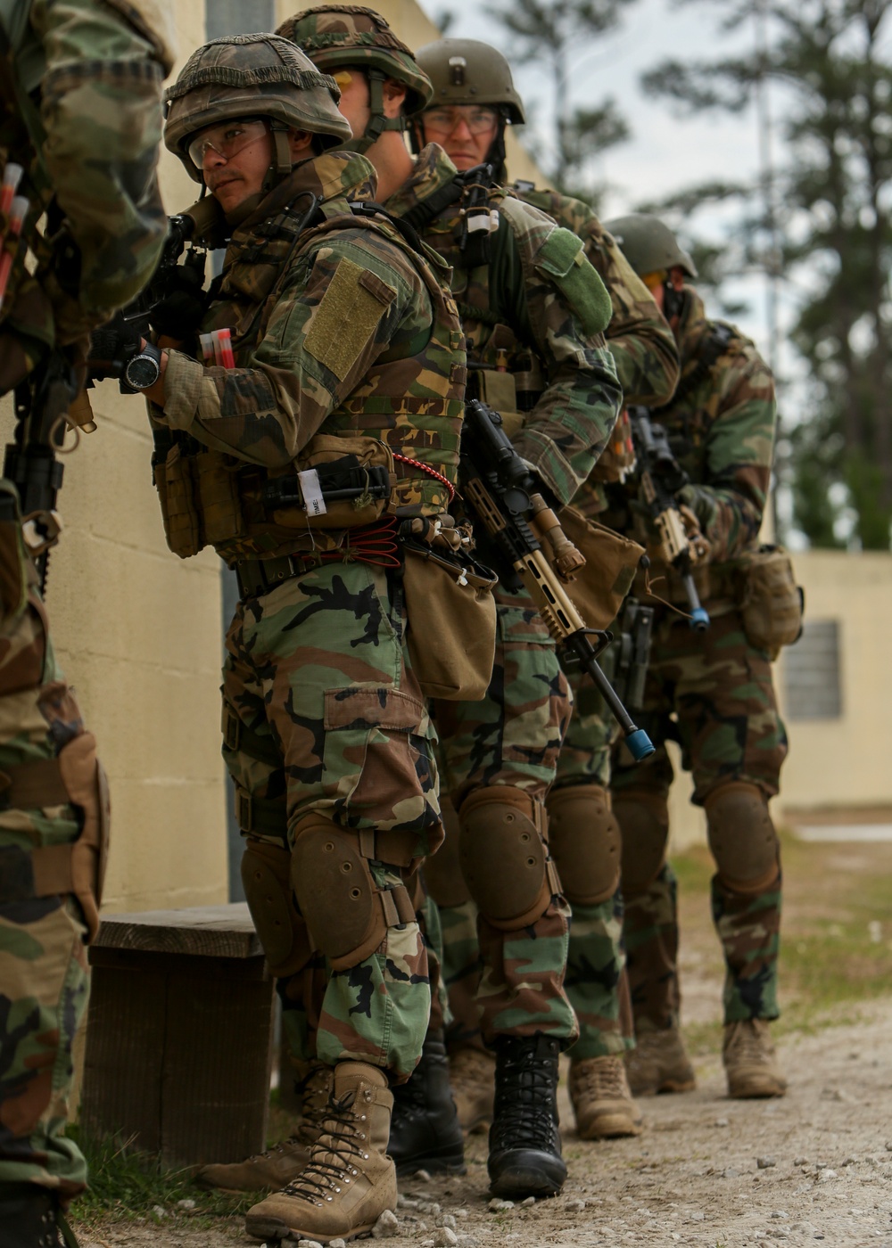 A group of military personnel in woodland camouflage (BDU pattern) are stacked against a building wall in a tactical formation, suggesting a military training exercise. The soldiers are fully equipped with helmets, body armor, weapons, and knee pads. Blue barrel caps on some weapons indicate this is a training/simulation exercise rather than live combat.