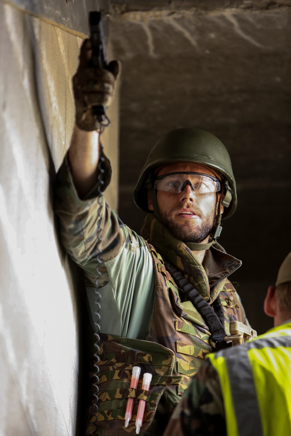 A male individual wearing military camouflage uniform and a green helmet with protective safety glasses is holding a tool or device up against a wall or ceiling junction inside a concrete structure. He appears to be conducting an inspection, measurement, or installation task. A second person in a high-visibility vest is partially visible in the background.