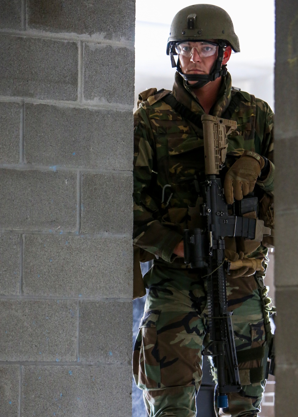 A soldier in woodland camouflage BDUs stands at the edge of a concrete block wall, holding a rifle in a ready position. The individual is wearing a PASGT-style helmet, clear protective eyewear, and tactical gloves, suggesting a training exercise or security operation. The setting appears to be a military training facility with cinder block construction.
