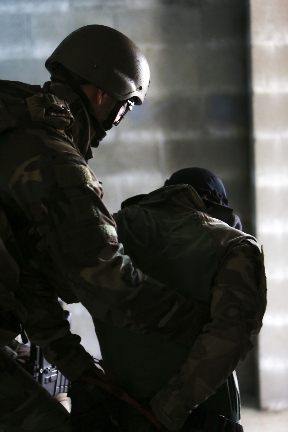 Two individuals in military/tactical gear are shown in close physical contact, with one appearing to restrain or guide the other. Both are wearing helmets and camouflage clothing, suggesting a military training exercise or tactical operation. The scene is set against a concrete or stone wall background in low-light conditions.
