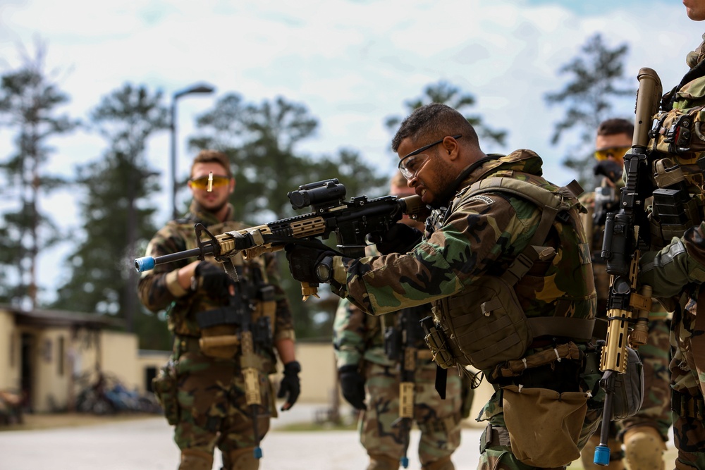 Military personnel in woodland camouflage uniforms are conducting a training exercise, likely force-on-force or simunition training based on the blue training barrel tips visible on the weapons. The central figure is aiming a rifle equipped with an optic and a blue-tipped training barrel, wearing protective eyewear and a tactical vest. Multiple personnel are visible in the background similarly equipped, suggesting a structured tactical training drill at a military installation.