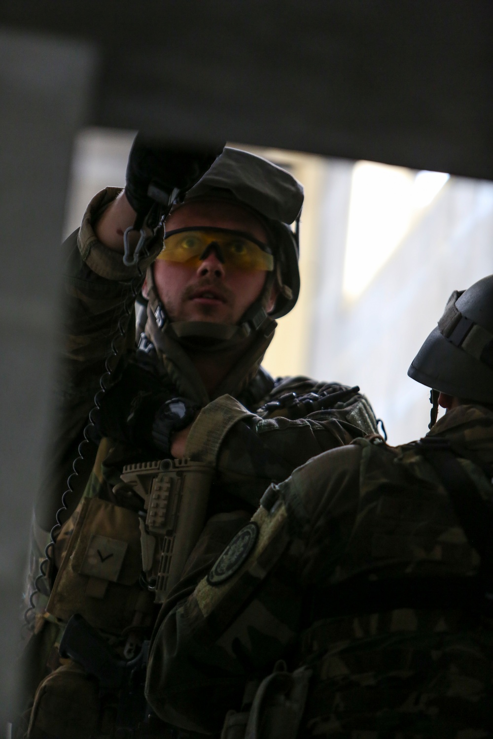 The image shows a military or tactical operator in full combat gear, appearing to be inside a building or structure. The primary subject is looking upward while holding what appears to be a coiled cable or cord near his helmet. A second helmeted individual is partially visible in the foreground right.