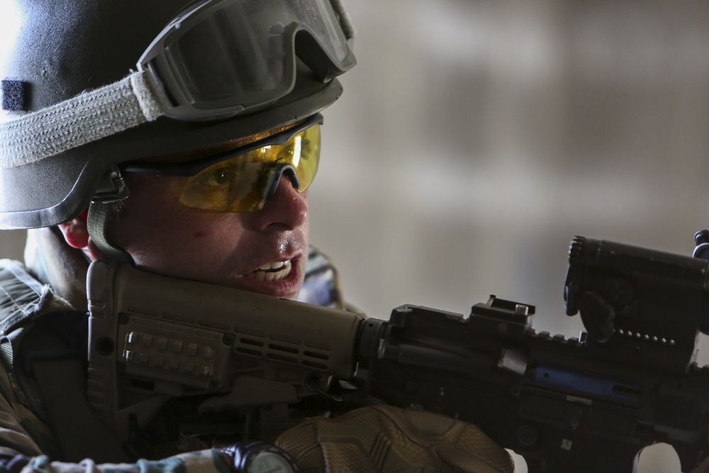 A close-up photograph of a military soldier in full tactical gear aiming a rifle. The soldier is wearing a ballistic helmet with mounted goggles, yellow-tinted protective eyewear, and body armor. The image is tightly framed, emphasizing the soldier's focused expression and weapon.