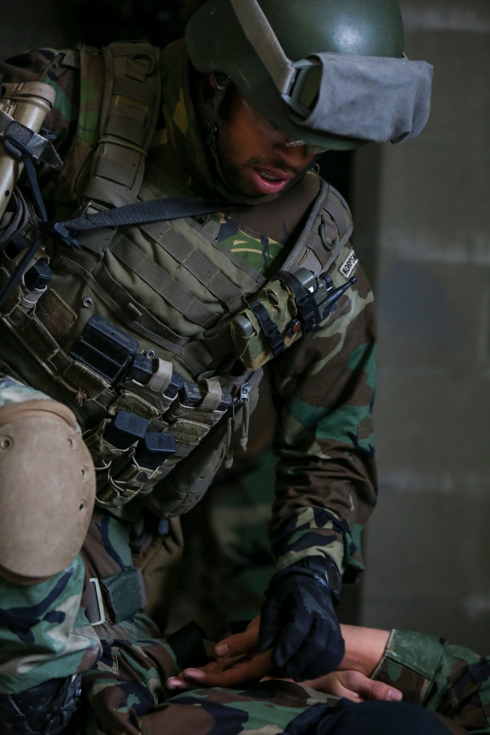 A soldier in full tactical gear, including a green PASGT-style helmet with attached goggles, plate carrier vest, and woodland camouflage BDU, is leaning over and attending to another person on the ground. The standing soldier appears to be performing a medical assessment or casualty care procedure. A partial name tape is visible on the vest, reading 'KORS...'