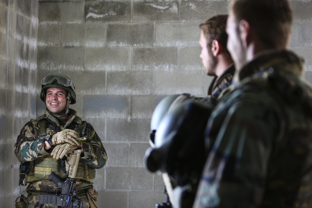 Three military personnel in camouflage uniforms are gathered inside a concrete block structure, appearing to be in a briefing or casual conversation. The soldier on the left is smiling and appears to be speaking or gesturing, while two others face him from the right foreground. The setting suggests a training exercise or operational staging area.