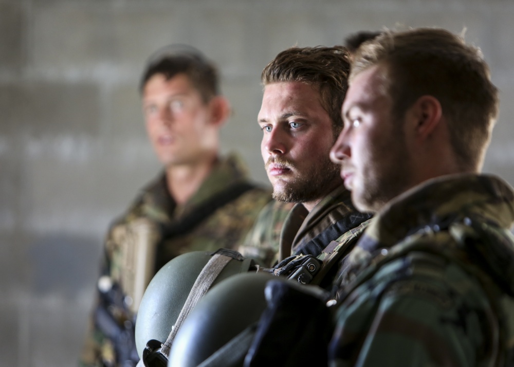 Three male soldiers in camouflage military uniforms are positioned in a row, appearing to be at attention or listening to a briefing. The central figure is in sharp focus, holding a military helmet, with a serious and attentive expression. The background shows a concrete wall structure, suggesting an indoor or semi-enclosed military facility.
