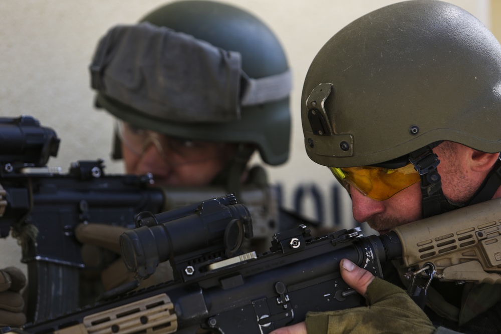 Two armed personnel in tactical gear are shown aiming rifles, with the foreground figure in sharp focus and the background figure slightly blurred. Both wear ballistic helmets and appear to be in a training or operational posture. The foreground individual wears amber-tinted protective eyewear and is sighting down a rifle equipped with an optic.