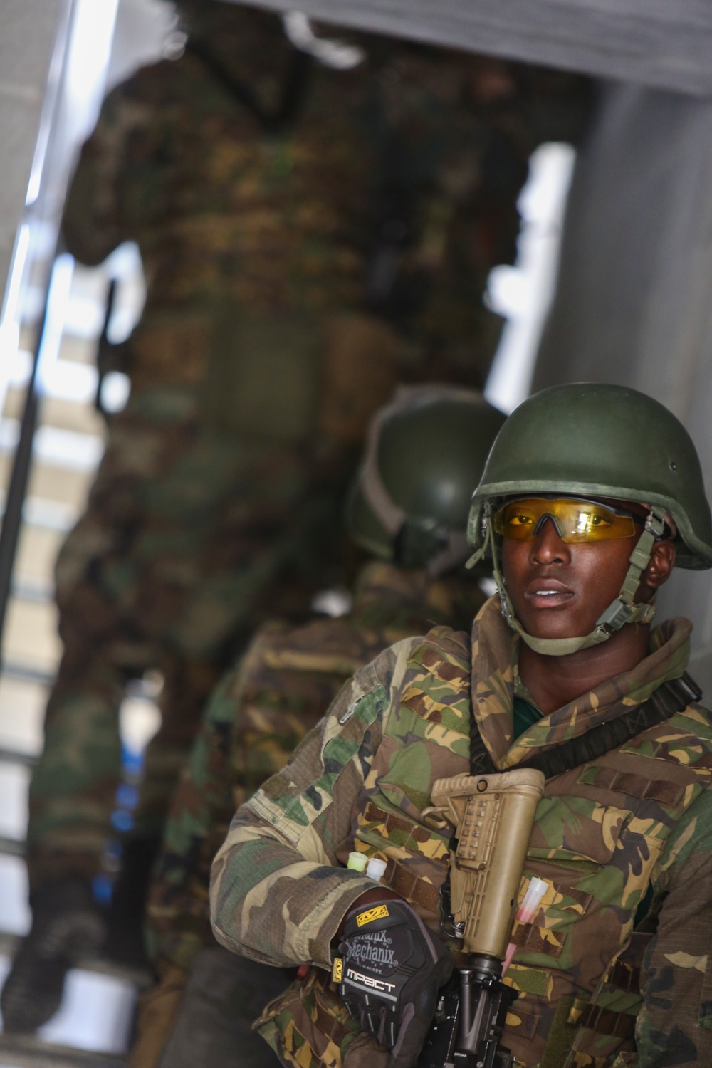 A soldier in the foreground is equipped with full combat gear including a green ballistic helmet, yellow-tinted protective eyewear, camouflage uniform, Mechanix Impact tactical gloves, and is holding a rifle. Behind him, additional soldiers in similar camouflage uniforms are visible but out of focus, suggesting a group formation or queue. The setting appears to be inside or near a military transport vehicle or structure.