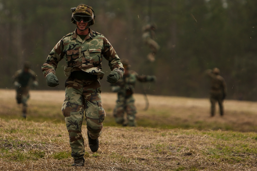 A soldier in woodland camouflage BDUs is running toward the camera across an open grassy field, smiling, while several other similarly uniformed personnel are visible in the blurred background. The setting appears to be a military training exercise or field operation in a wooded area. Debris or particles are visible in the air, suggesting recent physical activity or an explosion nearby.