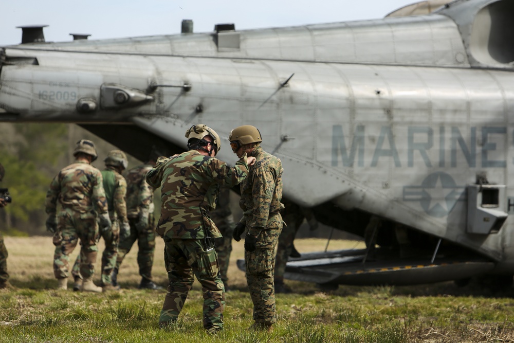 The image shows military personnel gathered near a large helicopter marked 'MARINES' with a visible tail number 162009, consistent with a CH-46 Sea Knight or similar tandem-rotor aircraft. Two soldiers in the foreground appear to be in close conversation or conducting an equipment check, while additional personnel are visible in the background. The setting appears to be an open grassy field, suggesting a training exercise or field operation.