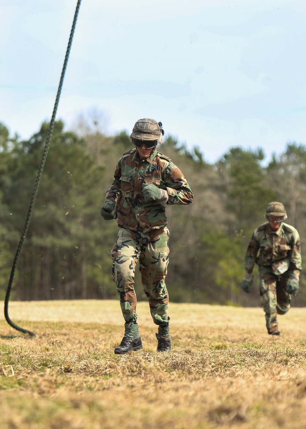 Two military personnel in woodland camouflage uniforms are running across an open grassy field during what appears to be a training exercise. The foreground soldier wears protective goggles, a boonie hat, and gloves, with a thick rope visible to their left. The setting is an outdoor training area bordered by a tree line under a clear sky.