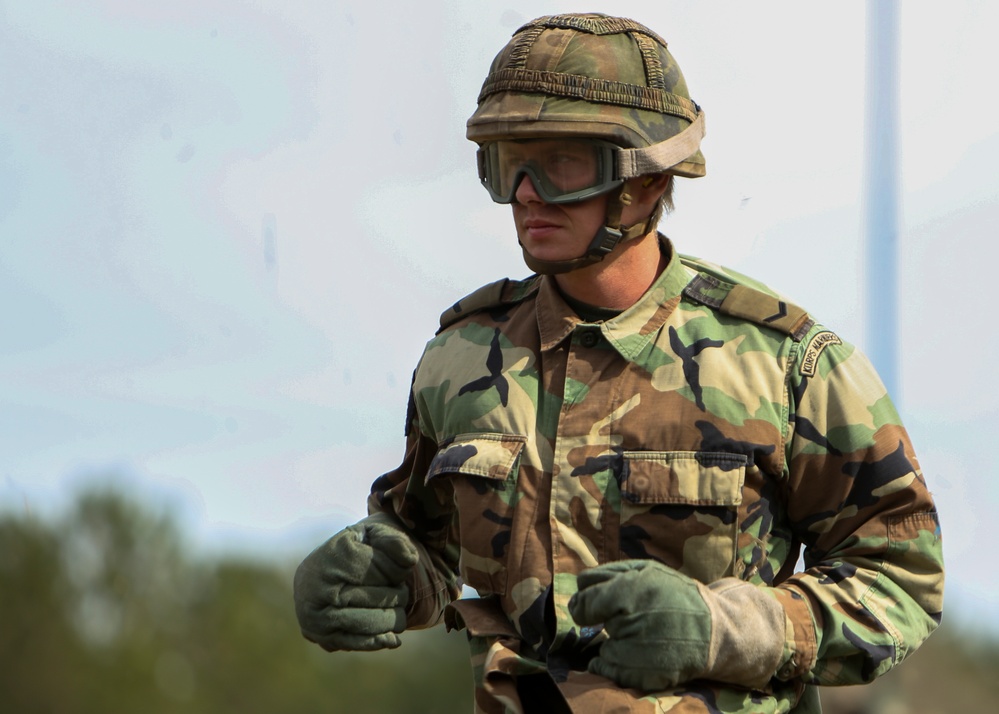 A male military service member is photographed in an outdoor setting wearing woodland camouflage battle dress uniform (BDU). He is equipped with a PASGT-style helmet with camouflage cover, ballistic goggles, and olive drab gloves. A branch tape or unit patch is visible on his right shoulder, consistent with U.S. Marine Corps insignia.