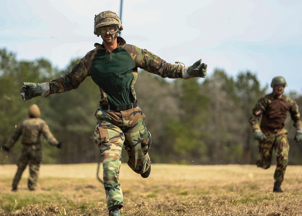 Three individuals in military camouflage uniforms are running across an open grassy field during what appears to be a training exercise. The foreground subject is in sharp focus, wearing woodland camouflage, a helmet, protective goggles, and gloves, with arms outstretched mid-stride. Two additional personnel are visible in the background, similarly equipped.