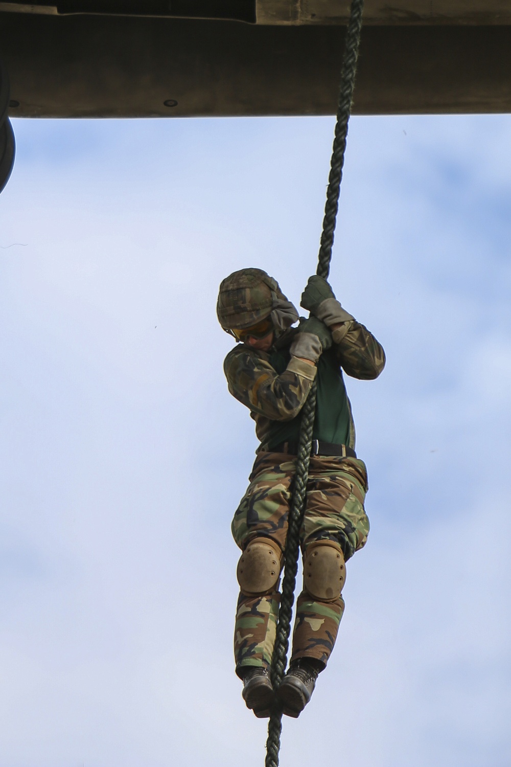 A soldier in camouflage uniform is climbing a thick rope suspended from an elevated wooden platform structure. The photo is taken from a low angle looking upward, capturing the soldier mid-climb against a partly cloudy sky. This appears to be a military training exercise or obstacle course activity.