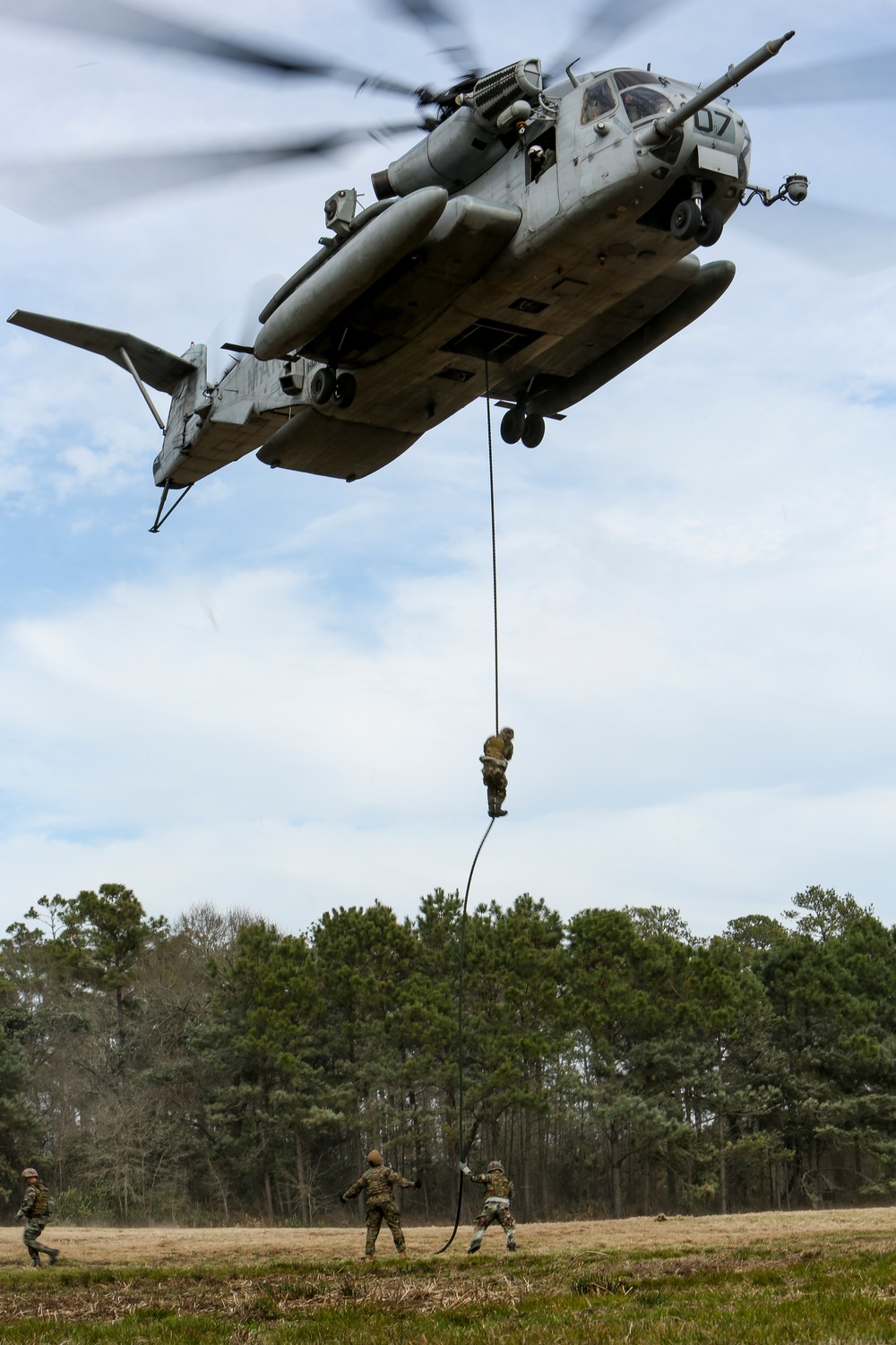 A large military helicopter, identifiable as a CH-53E Super Stallion marked '07', hovers over an open field while personnel conduct fast-rope or rappelling training. One soldier is visible mid-rope descending from the aircraft, while two personnel on the ground manage the rope below. The setting appears to be a military training area surrounded by pine trees.