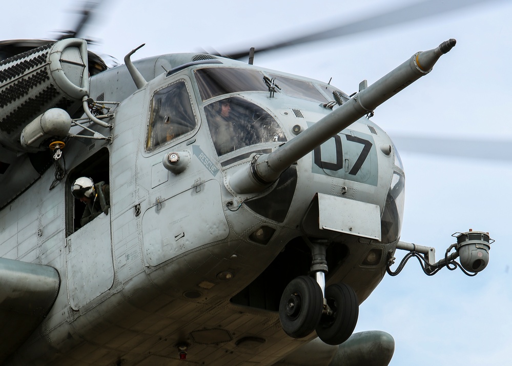 A close-up photograph of a military helicopter, identifiable by the number '07' on its fuselage and a 'RESCUE' label near the cockpit. The helicopter appears to be in flight or hovering, with rotor blades visibly in motion. At least two crew members are visible — one in the cockpit and one in a side window wearing a white helmet.