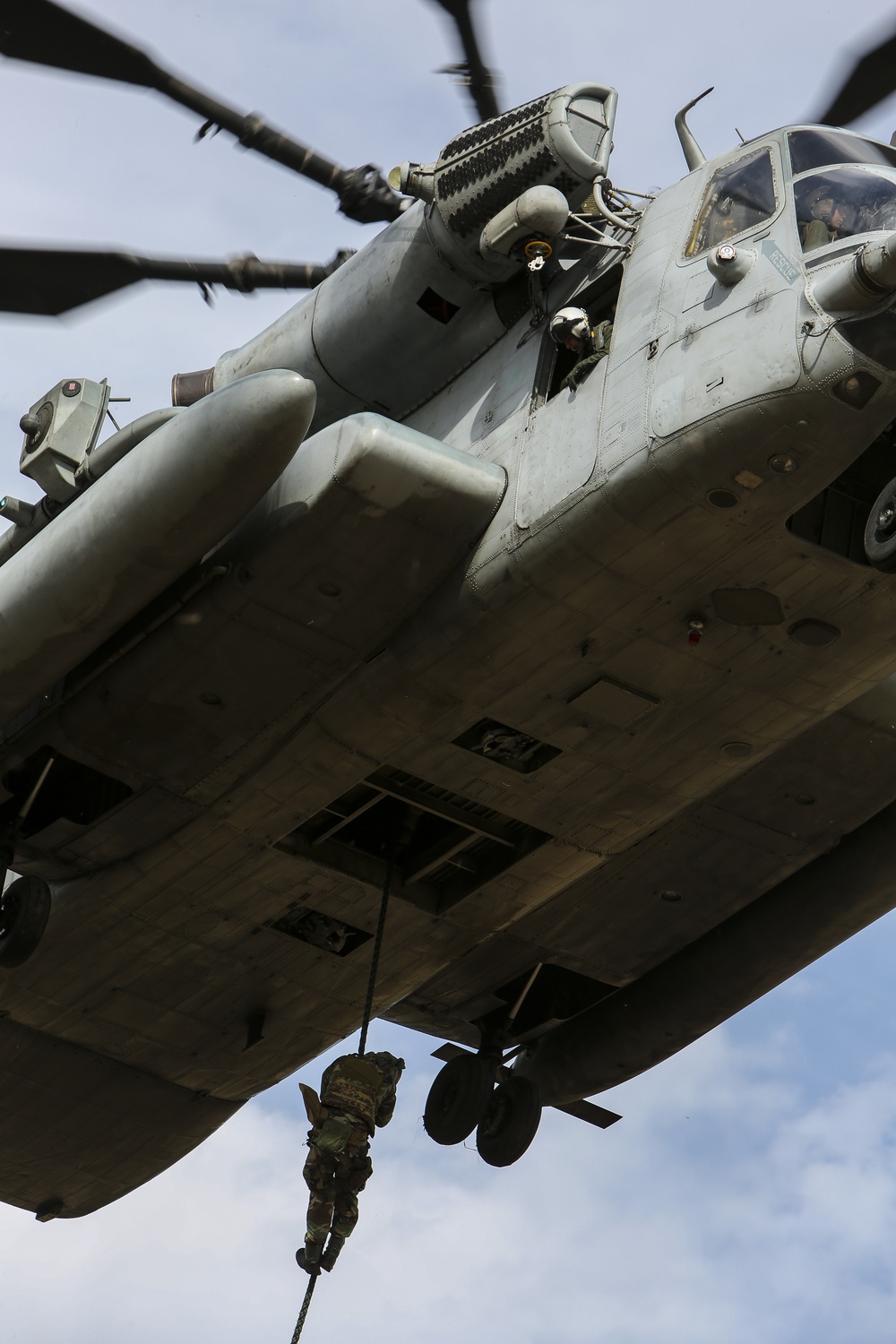 A large military helicopter, identifiable as a CH-53E Super Stallion, is photographed from below while in flight. A soldier in camouflage gear is rappelling or fast-roping down from the aircraft's underside via a rope. At least one crew member is visible in the cockpit and another appears to be observing from an open door.