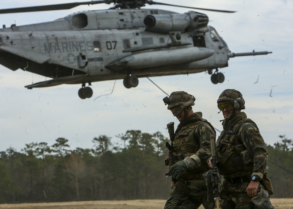 Two U.S. Marines in full combat gear walk across an open field as a large CH-53 Sea Stallion helicopter marked 'MARINES 07' hovers low overhead. Debris and vegetation are visibly disturbed by the rotor wash. The scene appears to be a military training exercise conducted in a wooded, open terrain environment.